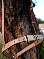 A Rawhide with Turquoise Studs- Sliding One Ear is hanging on a tree trunk. The woven section features a natural straw color with black accents, and the leather parts are a combination of light and medium brown. Background includes grass and partial sky.