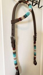 A One Ear Headstall - Teal Serape decorated with colorful, striped woven bands in teal, white, brown, and red is hanging on a white wall. The headstall includes metal buckles and stitching details.