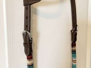 A One Ear Headstall - Teal Serape decorated with colorful, striped woven bands in teal, white, brown, and red is hanging on a white wall. The headstall includes metal buckles and stitching details.