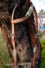 A Rawhide with Turquoise Studs- Sliding One Ear is hanging on a tree trunk. The bridle features metal buckles and is set against an outdoor backdrop, including a blurred fence, yard, and garden tools.