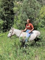 A person with long hair, wearing a brown T-shirt and blue jeans, rides a white horse adorned with a Turquoise Breeze - Hand Tooled/Painted - Sliding One Ear Headstall through a lush, green forest. The area is abundant with tall grass, bushes, and trees, with hints of sunlight breaking through the foliage. The person is smiling and relaxed.