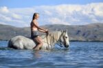 A young woman with braided hair and glasses rides a white horse adorned with a Turquoise Breeze- Hand Tooled/Painted- Sliding One Ear Headstall through shallow water. She is wearing a red and white patterned bikini top and gray shorts. The background features hills and a partly cloudy sky.