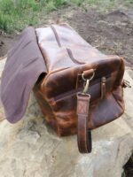 A Distressed Leather Duffel Bag / Travel Bag- The Vintage is sitting on a large rock. The bag has a zipper pocket on the side, a strap, and brass hardware. The background shows a grassy and dirt area.