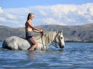 A woman wearing a bikini top and shorts rides a white horse through water, partially submerged. The horse sports a Turquoise Breeze- Hand Tooled/Painted- Browband Headstall. In the background are hills and a partly cloudy sky. The scene is bright and sunny, suggesting a warm, pleasant day.