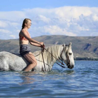 A woman wearing a bikini top and shorts rides a white horse through water, partially submerged. The horse sports a Turquoise Breeze- Hand Tooled/Painted- Browband Headstall. In the background are hills and a partly cloudy sky. The scene is bright and sunny, suggesting a warm, pleasant day.