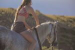 A person with a long braid, wearing a striped bikini top and patterned shorts, is riding a white horse in a hilly landscape. The horse is adorned with a Turquoise Breeze- Hand Tooled/Painted- Browband Headstall. The backdrop features grassy terrain and a light blue sky.