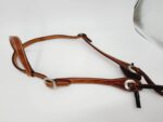 A Cowpuncher Sliding One Ear Headstall With Tooling with silver buckles and detailed stitching, featuring a belt style headstall, is laid out on a white surface. A reflective silver object is partially visible in the background.