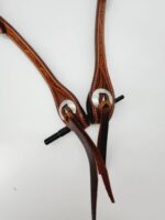 A close-up of intricately tooled brown leather straps with silver buckles and long leather strings attached, resembling a Cowpuncher Sliding One Ear Headstall With Tooling. The straps feature detailed stitching and decorative patterns along their length, set against a plain white background.