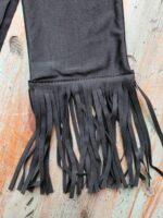 A piece of black fabric with a fringe trim lies on a rustic wooden surface, resembling the textured designs of Tail Bags. The fringe consists of multiple strips of fabric hanging down, creating a decorative effect that contrasts with the distressed, light-colored wood background.