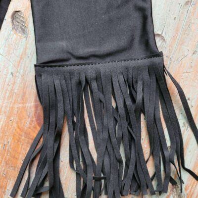 A piece of black fabric with a fringe trim lies on a rustic wooden surface, resembling the textured designs of Tail Bags. The fringe consists of multiple strips of fabric hanging down, creating a decorative effect that contrasts with the distressed, light-colored wood background.