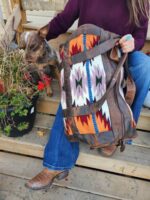 A person wearing blue jeans and cowboy boots holds a colorful, patterned Leather Saddle Blanket Duffel Bag- Tribal Sunset while sitting on wooden steps. A dog with a brown and white coat sits beside them, sniffing a pot of plants with red flowers. The person wears a maroon shirt and has turquoise jewelry.