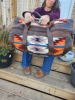 A woman holding a large Leather Saddle Blanket Duffel Bag- Tribal Sunset with both hands. The bag features a colorful geometric design. She is seated on wooden steps, wearing a maroon top, blue jeans, brown boots, and turquoise rings. Potted plants are nearby.