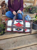 A person holding a Leather Saddle Blanket Duffel Bag- The Alpine with tribal designs sits on wooden steps. A dog is sitting beside them. Several pots of plants surround the area. The person wears a maroon top and blue jeans.