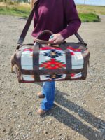 A person wearing a maroon sweater and blue jeans is holding a Leather Saddle Blanket Duffel Bag- The Alpine with tribal patterns in red, black, and white. The bag has brown leather straps and panels. They are standing on a gravel road with a picturesque countryside landscape in the background.