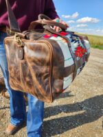 A person in jeans and a burgundy top holds a Leather Saddle Blanket Duffel Bag - The Alpine. The background shows a dirt path and a clear blue sky with scattered clouds.