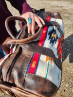 A close-up photo shows a hand with a turquoise ring resting on the handle of The Alpine. The bag features colorful, southwestern-style woven fabric, including red, blue, white, and black geometric patterns. The background is a gravel surface.