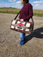 A person in a maroon long-sleeve shirt and blue jeans stands on a gravel path in a rural area, carrying the Leather Saddle Blanket Duffel Bag- The Alpine. The background reveals a grassy field and partly cloudy sky.