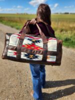 A person with long hair walks on a dirt path carrying a large Leather Saddle Blanket Duffel Bag- The Alpine with a colorful southwestern pattern. The bag has brown leather accents, and the person is wearing a dark jacket and jeans. In the background, a grassy field stretches under a clear blue sky.