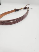 A close-up image of a brown leather belt with white stitching. The belt has a colorful beaded section near the buckle and brown leather laces at the end. The background is white and minimalistic.