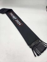 A black sash with white stars and "Trump 2024" printed in white and red letters. The Tail Bag- Trump 2024, reminiscent of mane bags, has several narrow black ribbons hanging from one end and is laid out on a light-colored surface.