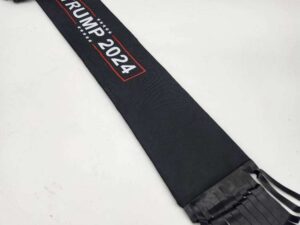 A black sash with white stars and "Trump 2024" printed in white and red letters. The Tail Bag- Trump 2024, reminiscent of mane bags, has several narrow black ribbons hanging from one end and is laid out on a light-colored surface.