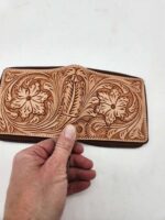 A hand holding a tan leather wallet that features intricate floral and swirl engraving designs, set against a plain white background. The artwork on the wallet is detailed and showcases skilled craftsmanship.