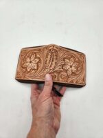 A hand is holding a brown leather wallet adorned with intricate floral and leaf patterns. The carving details are elaborate and cover nearly the entire visible surface of the wallet. The background is a plain white surface.