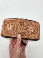 A hand holds an intricately crafted leather wallet. The wallet features detailed floral and leaf patterns, showcasing fine craftsmanship. The background is a plain, white surface.