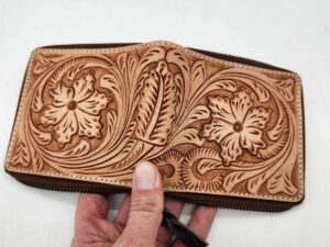 A hand holds an intricately crafted leather wallet. The wallet features detailed floral and leaf patterns, showcasing fine craftsmanship. The background is a plain, white surface.