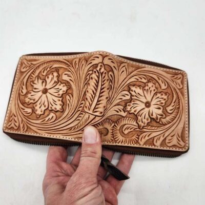 A hand holds an intricately crafted leather wallet. The wallet features detailed floral and leaf patterns, showcasing fine craftsmanship. The background is a plain, white surface.