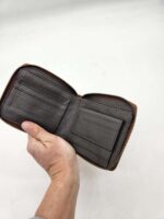 A hand is holding an open brown leather wallet with a zipper. The wallet displays several card slots and a section for a small wallet or coin pouch on the right side. The background is a plain white surface.