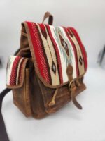 A brown leather backpack featuring a top flap with a red, white, and blue geometric woven fabric design. The backpack has a front pocket with a strap closure and matching fabric accents. The bag stands on a white surface against a neutral background.