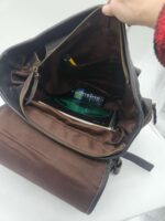 A person holds open a brown, insulated bag with a partially unzipped top. Inside the bag are a black pen, a small blue water bottle, a green plastic container, and a book. The bag has several pockets and compartments.