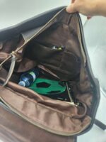 A person holds open a brown zippered bag showing its contents, which include a green and black item resembling a soccer ball notebook, a partially visible blue water bottle, and a few small items tucked into inner pockets. The bag has a dark brown interior lining.