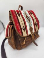 A brown leather backpack with a patterned flap featuring a red, white, and blue geometric design. The bag has leather straps and a side pocket with a matching pattern. The background is a plain surface, and the backpack is positioned at an angle.