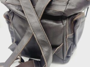 A dark brown leather backpack with adjustable straps laying crisscrossed on a white surface. The backpack features multiple pockets and a top flap closure. The leather has a textured appearance with visible stitching along the edges.