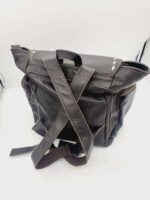 A black leather backpack is shown with beige stitching. It has shoulder straps crossed over each other and lying on the flat surface. The backpack features a rolled-up top and a handle for carrying. The background is white.