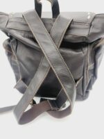 A dark brown leather backpack with light brown stitching is displayed. The backpack features adjustable straps and multiple pockets. The focus is on the back side, highlighting the crisscross pattern of the straps. The background is white.