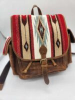 A brown leather backpack with a flap featuring a colorful woven design in red, beige, white, black, and blue patterns. The design includes geometric shapes and arrows. The bag has side pockets and a handle at the top.