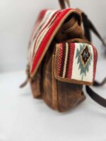 Side view of a rustic brown leather backpack with a woven red, white, and green patterned flap and side pocket. The bag has a vintage look with visible stitching and straps. The fabric design features geometric shapes and adds a Southwest style.