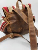 A rustic brown leather backpack with a southwestern-patterned fabric trim is shown. The fabric features red, white, and blue stripes. The bag has a buckle strap closure and adjustable shoulder straps, with a carry handle at the top.