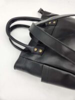 A close-up photo of a black leather handbag with two sturdy handles and brass rivets. The bag is set against a white background, showcasing its smooth texture and durable straps.