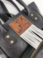 Close-up image of a black leather handbag featuring a brown leather patch with a floral design and white fringe details. The bag has brass rivets and a visible zipper. The texture and craftsmanship of the bag are clearly visible.