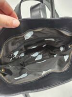 Close-up view of a black leather handbag with a hand partially opening the zippered main compartment. The inside lining of the bag is gray with white polka dots, and includes a small inner zippered pocket.