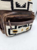 Close-up of a backpack featuring patterned fabric and brown leather accents. The bag has a front pocket with a zipper partially open, revealing a glimpse of the brown leather interior. The fabric pattern includes black, white, and yellow geometric shapes.