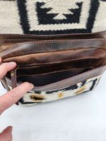 A brown leather and wool bag featuring a black and white geometric pattern is open, revealing multiple compartments and pockets. A person's hand is opening one of the zippered compartments. The bag is resting on a white surface.