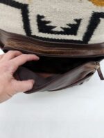 A hand is opening the pocket of a brown leather bag with a zipper. The interior of the pocket is empty. The outer part of the bag has a textile pattern with black and yellow designs.