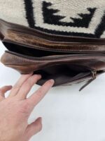 A hand is pulling open the zipper of a brown leather bag with a white and black woven fabric panel. The woven fabric displays a geometric pattern. The interior of the bag is visible, showing a dark lining.