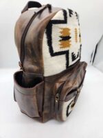 A brown leather backpack with a front section that features a white fabric with black and gold geometric patterns. The backpack has multiple compartments and gold zippers. The side pocket appears open, and the bag is set against a plain white background.