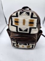 A brown leather backpack with a large front pocket and flap featuring a white, black, and gold geometric pattern. The bag is positioned upright against a white backdrop.